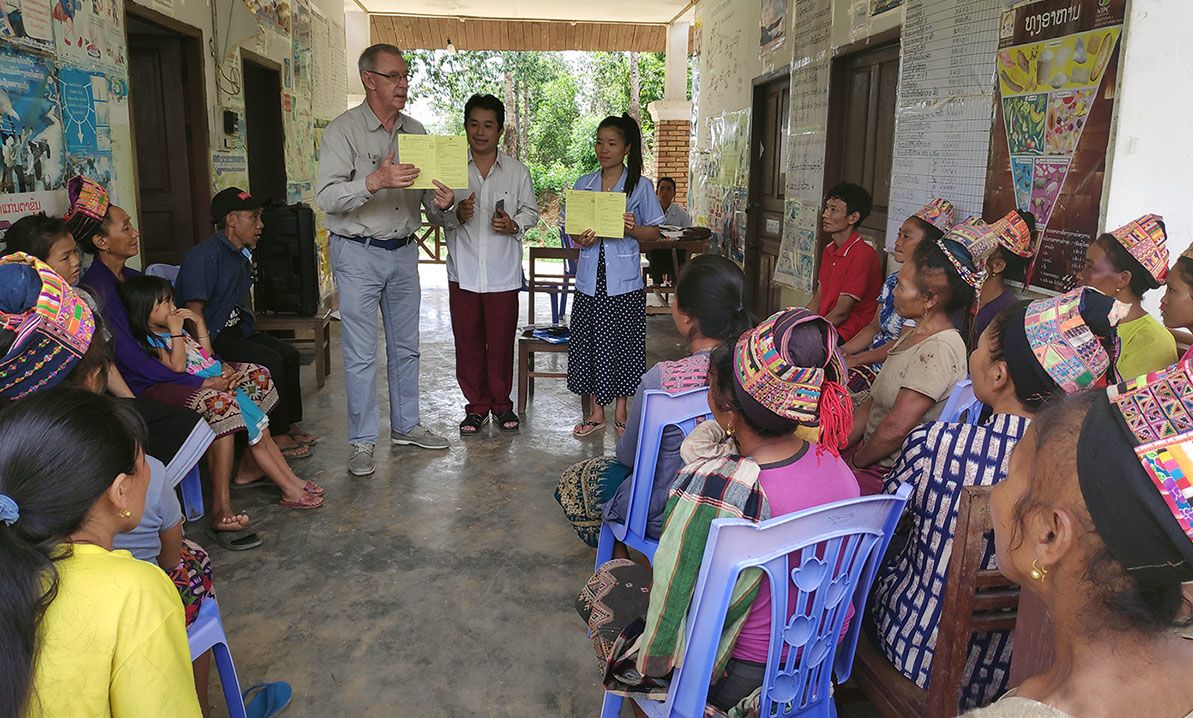 Julian holding a yellow vaccination card, speaking to a group of women in Laos