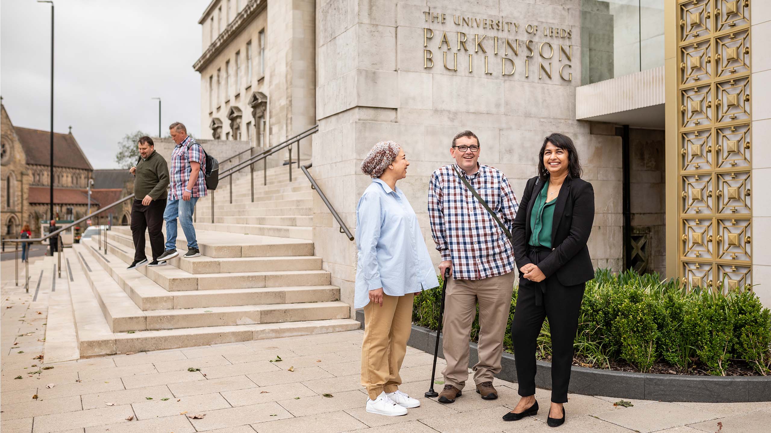 Three adults stand in front of the Parkinson Building as two adults walk down some steps behind them.