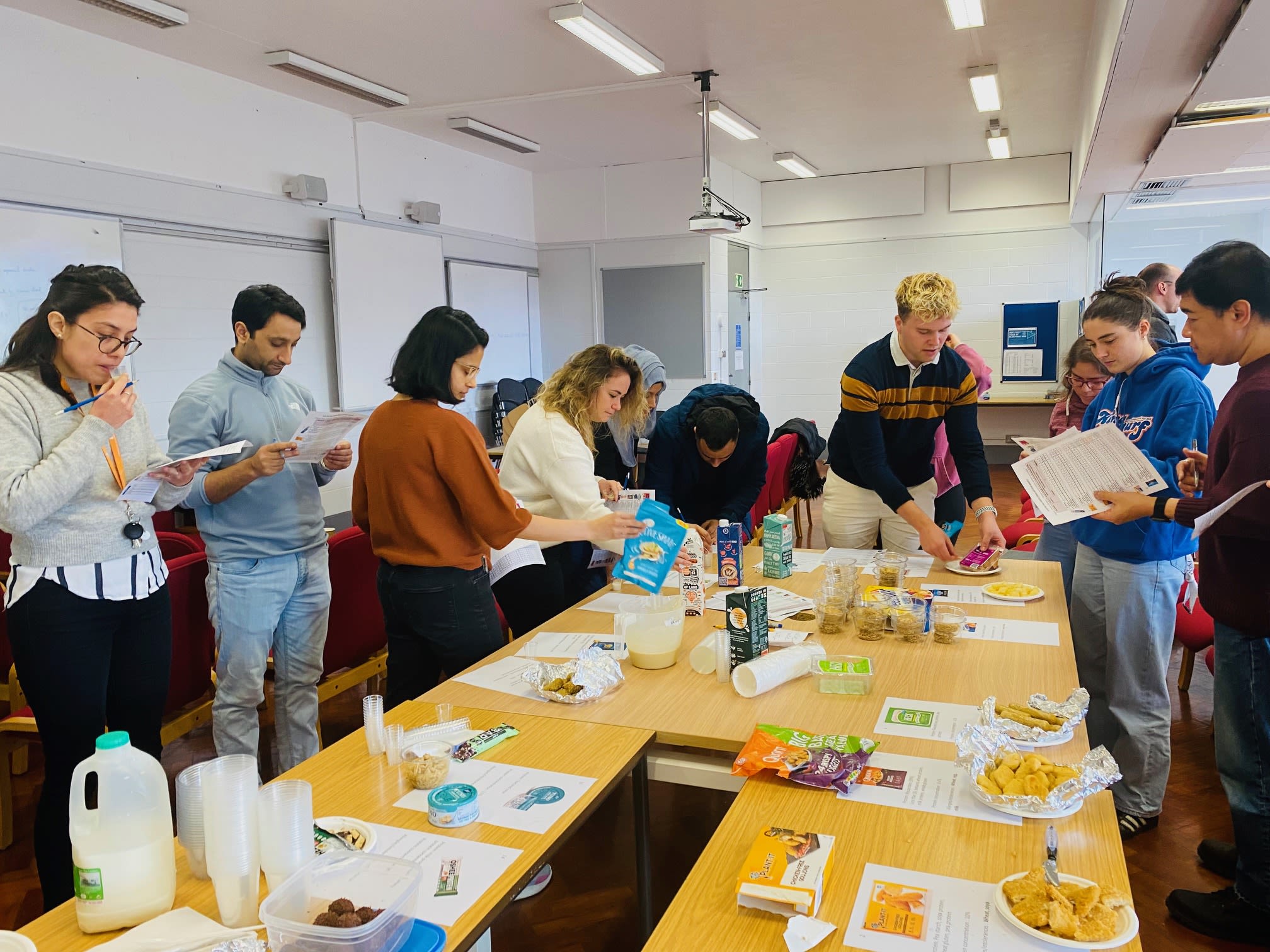 A group of students taste foods around a large table in a classroom environment, making notes on the textures as they experience them.