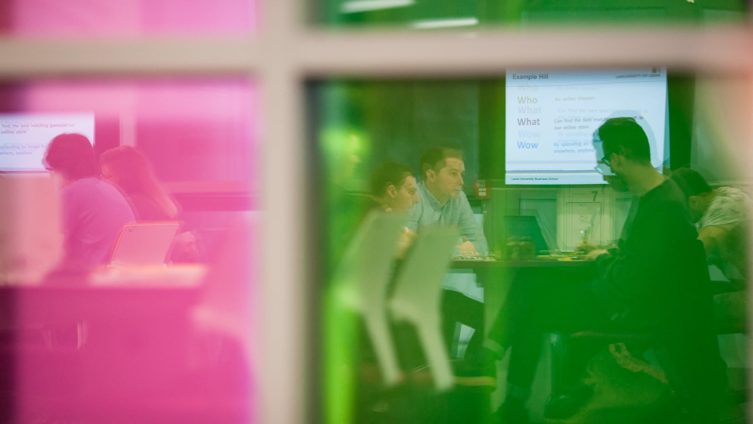 Two small groups of people sat in a teaching space chatting and using screens showing presentations