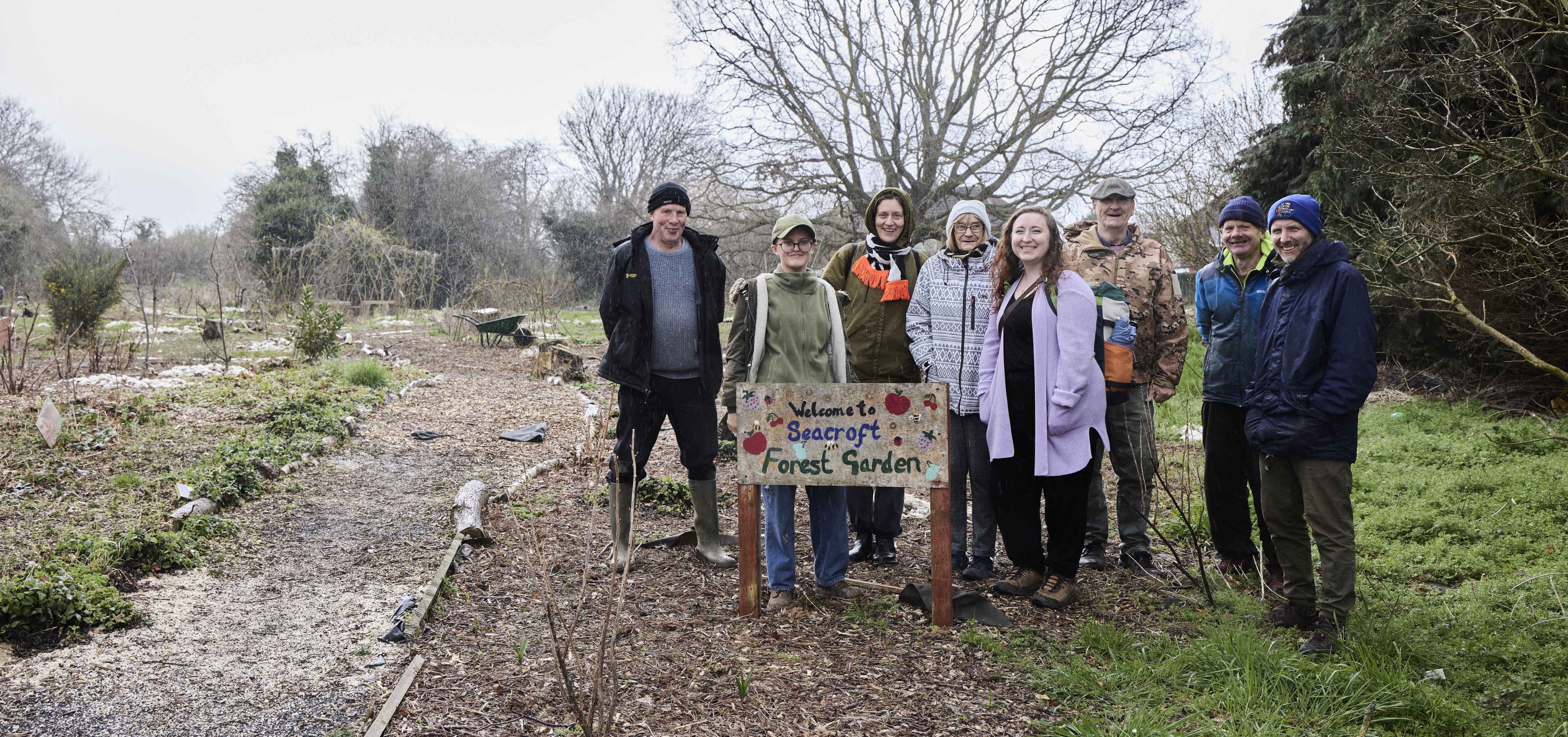 A group of seven people standing in a garden, with a hand-painted sign saying 'Welcome to Seacroft Forest Garden'. There's a path into the garden and trees in the background.