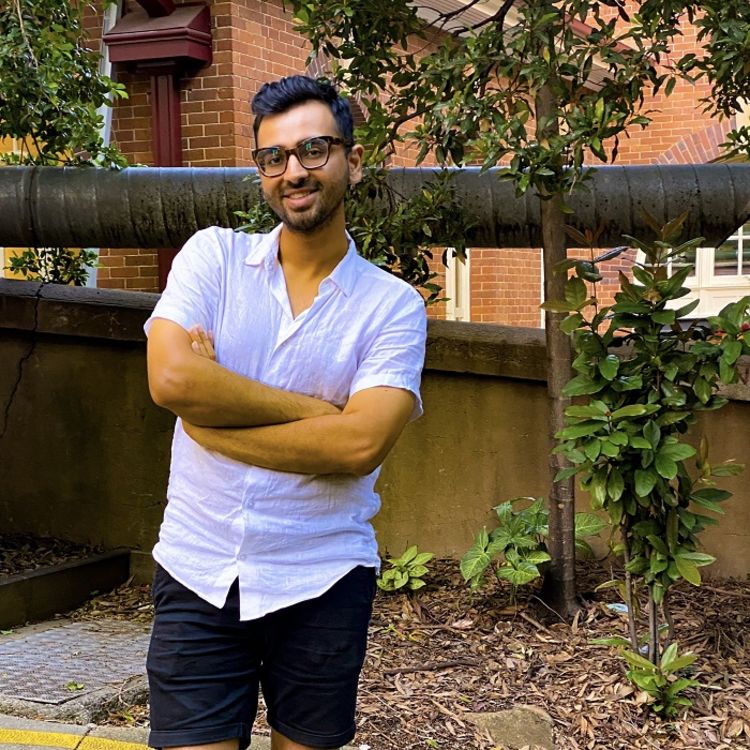 Photo of Rahul SInha-Roy stood in front of a tree wearing a white shirt. He is smiling and has his arms crossed.