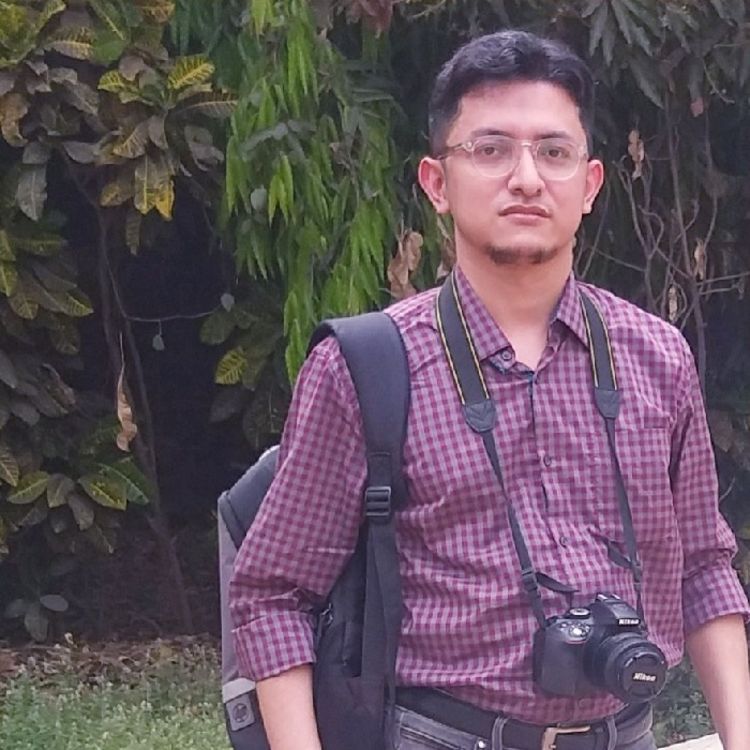 Photo of Arijit Das. He is wearing a purple checked shirt, with a bag hanging from his shoulder and a camera hung around his neck.