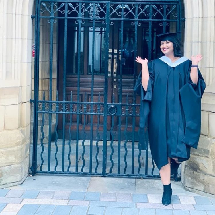 Photo of Ramya Baskaran in her graduation cap and gown, posing in front of the gates to the great hall at the university of leeds.
