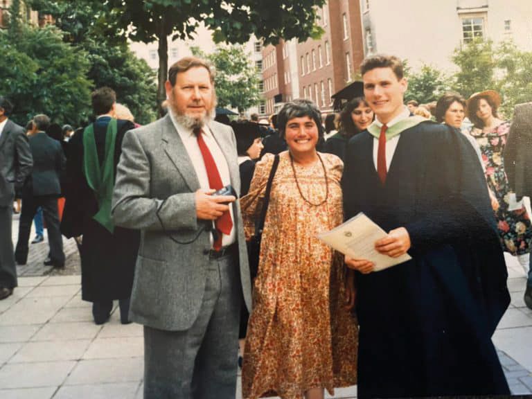 Keir on graduation day, in graduation gown and holding his degree certificate, with his parents outside The Great Hall