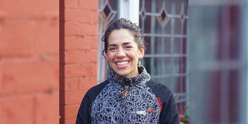 Rosie Vohra, artist, smiles. A red-painted brick building is behind her.
