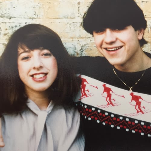 An old photo of a couple from the 1980s. The woman on the left has long dark hair and is wearing a grey blouse. The man on the right wears a woolen jumper with skiers on and a gold necklace.