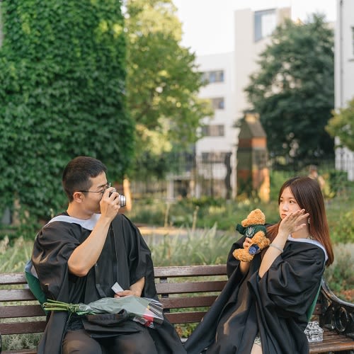 Picture of a couple on their graduation day sat on a bench. The man on the left takes a photo of a woman on the right
