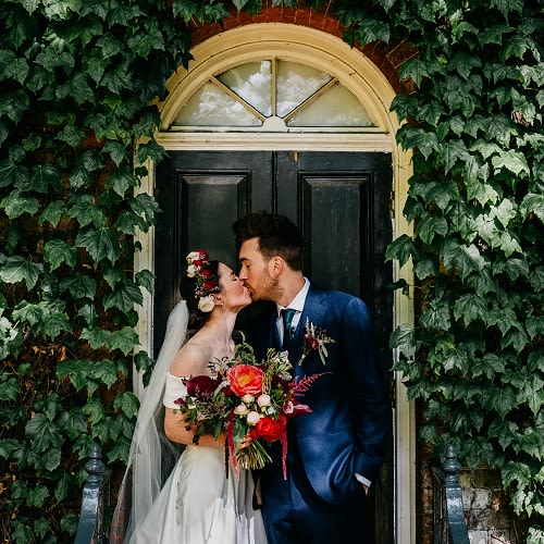 A couple on their wedding day kissing in front of a black door. The door is framed by ivy