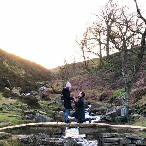 Photo of a man on one knee proposing to a woman on a bridge