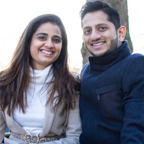 A couple posing or a photo smiling at the camera. The man on the right wears a blue coat, the woman on the left wears a white blouse and grey cardigan