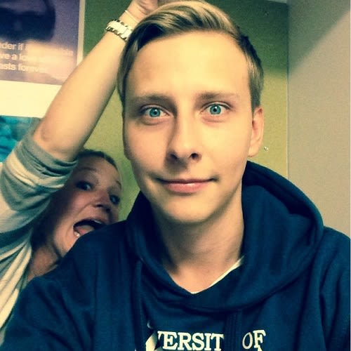 A portrait photo of a man wearing a uni of leeds hoodie. A woman is pulling a face behind him