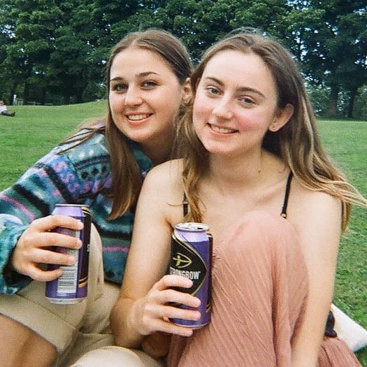 Two women smiling for the camera, sitting on some grass and both holding cans of Strongbow Dark Fruits