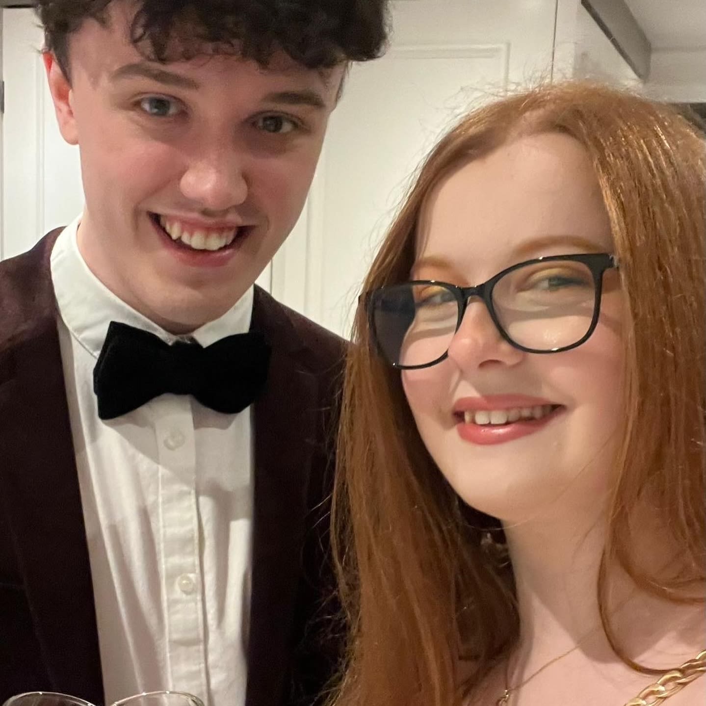A man and a woman smiling for the camera. The man is wearing a suit and bowtie and the woman is wearing thick rimmed glasses