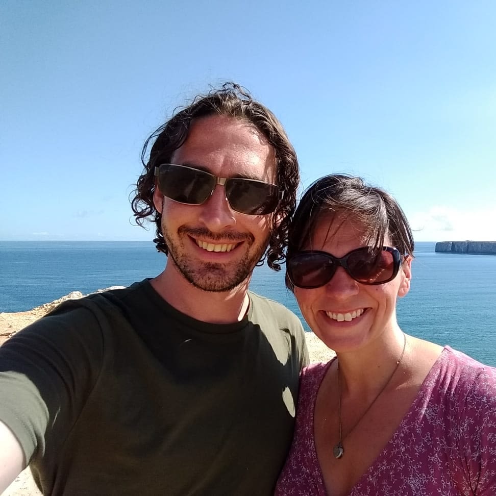A man and a woman pose for a photo in front of the sea. Both are wearing sunglasses and standing in the sun