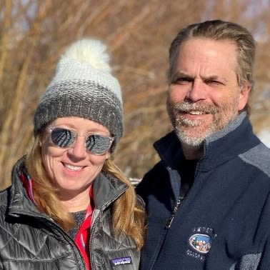 A man and a woman smiling for a photo. The woman is wearing sunglasses and a wooly hat