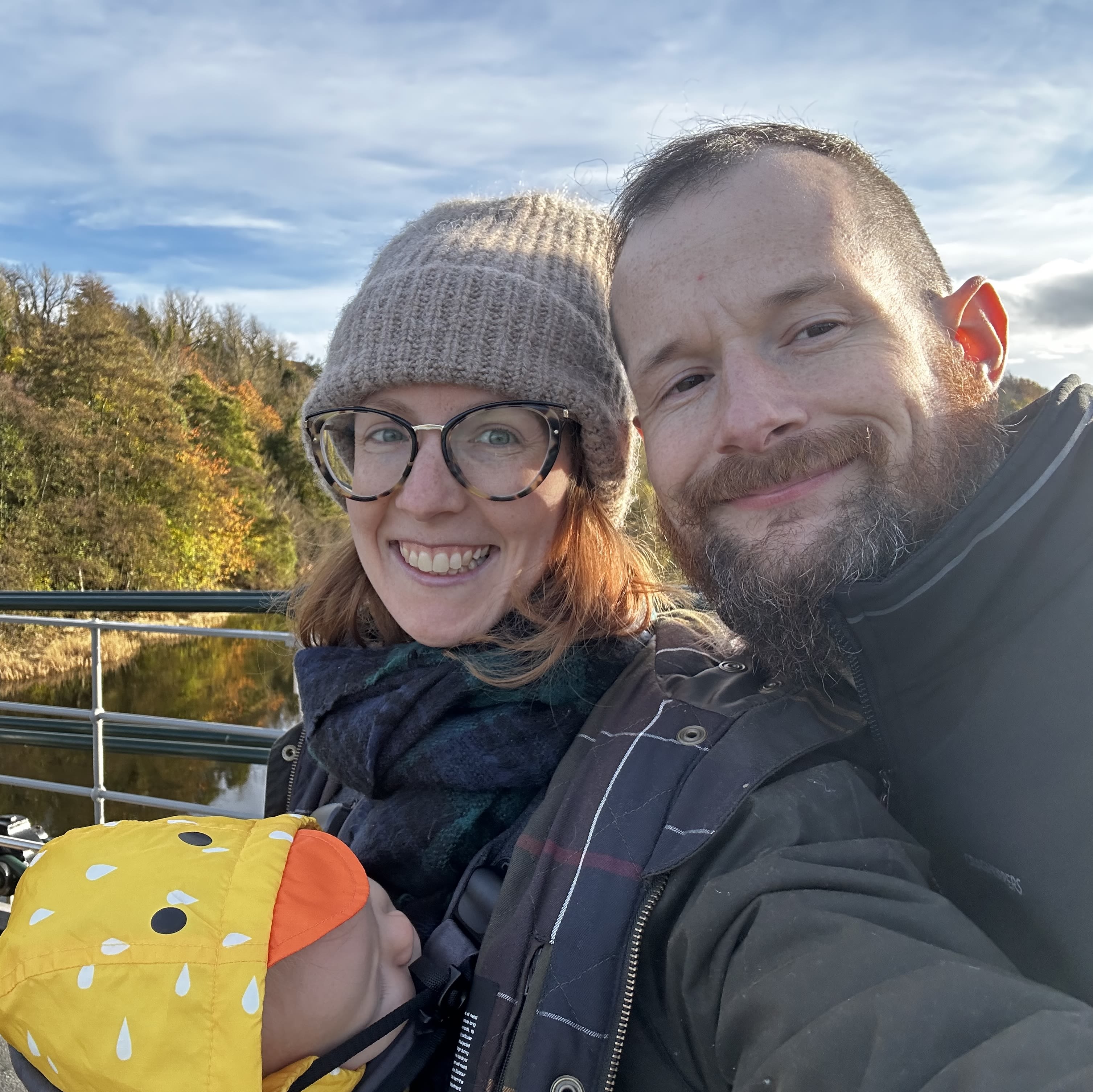 A man and a woman stand on a bridge smiling at the camera. The woman had a baby attached to her.