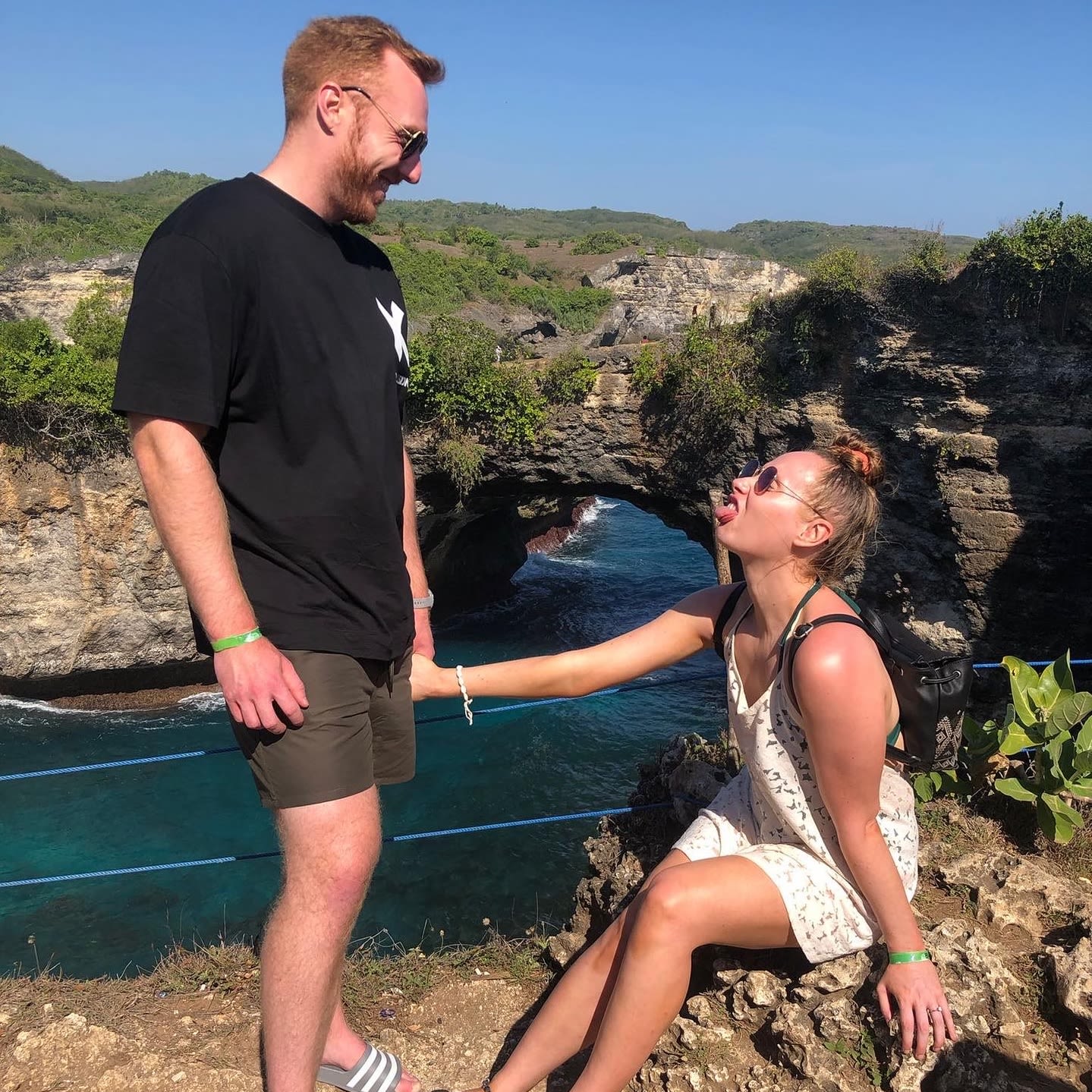 A man and a woman on a clifftop above the sea. The man looks at the woman sat on the floor, she has her tongue poking out