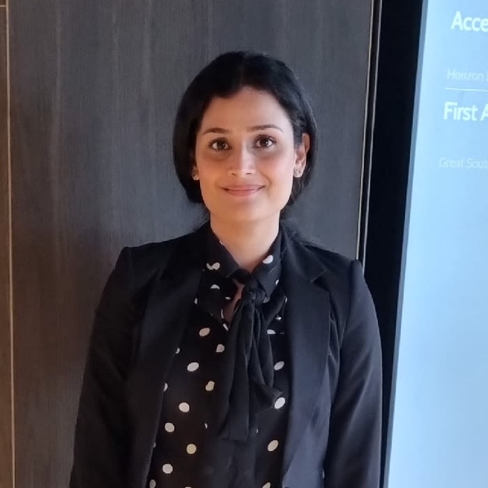 Photo of Hira Ali stood in front of a screen, wearing a polkadot blouse and black blazer