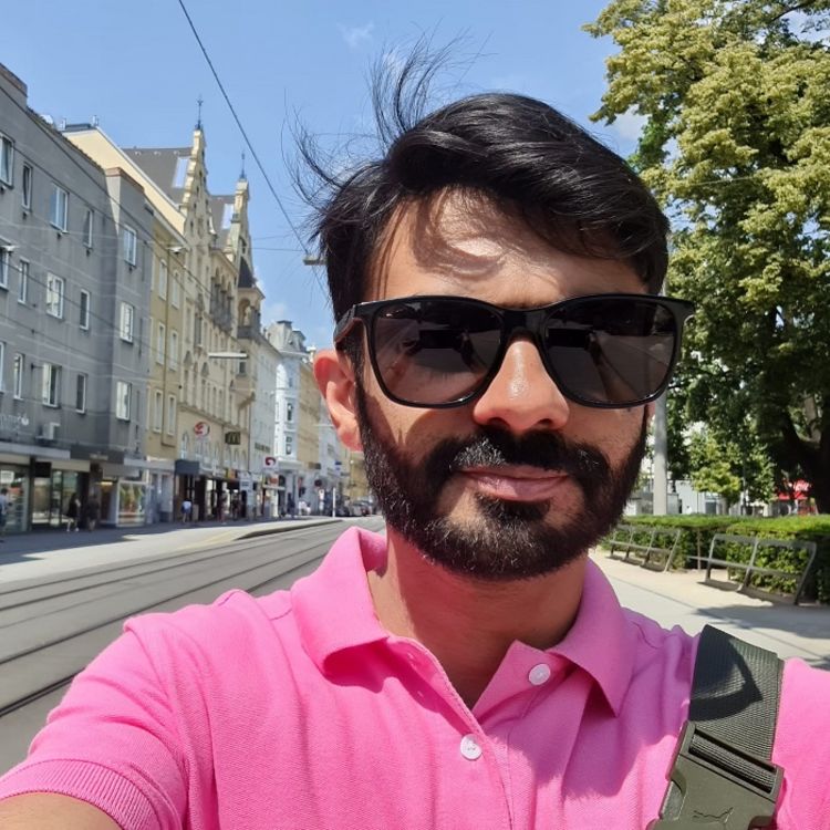 Selfie of Aitsam Hasan walking down a street wearing sunglasses and a pink t-shirt