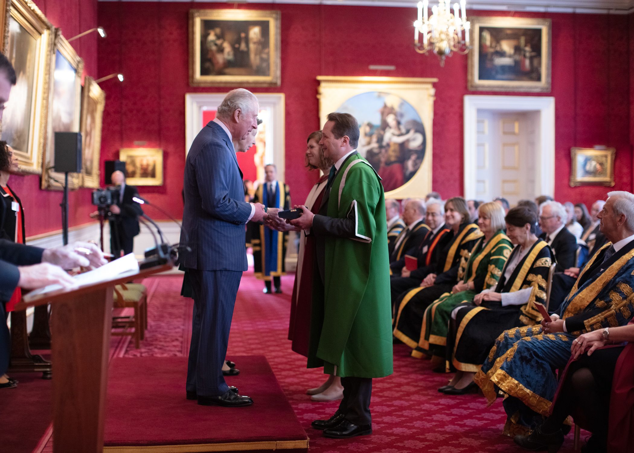 Nick Plant and Amanda Maycock stand at the front of the ceremonial room and Prince Charles and Princess Anne present them with the Queen's Anniversary Prize