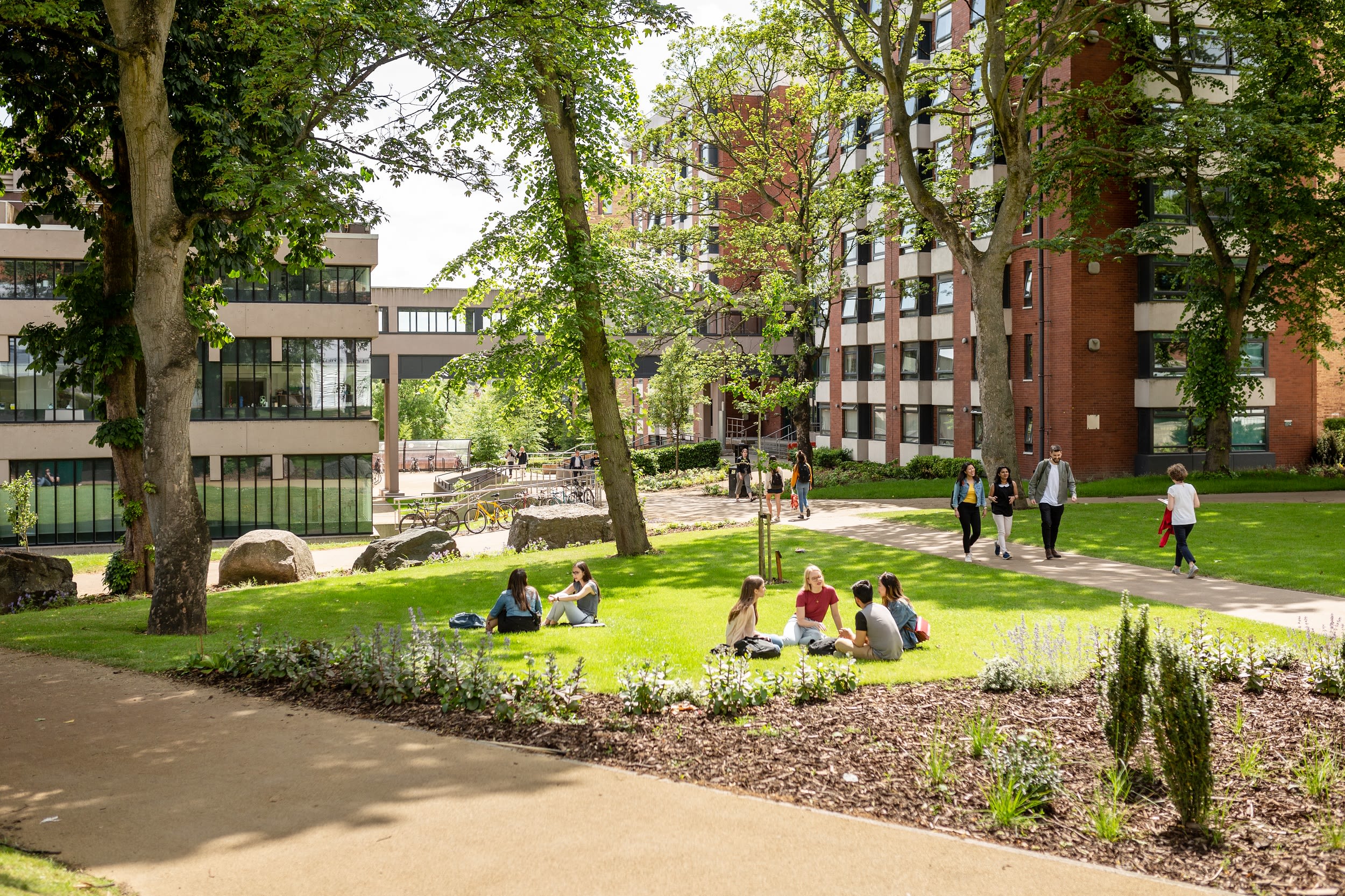 Two groups of students sat on grass at the University of Leeds campus on a sunny day with various students walking past on the path.