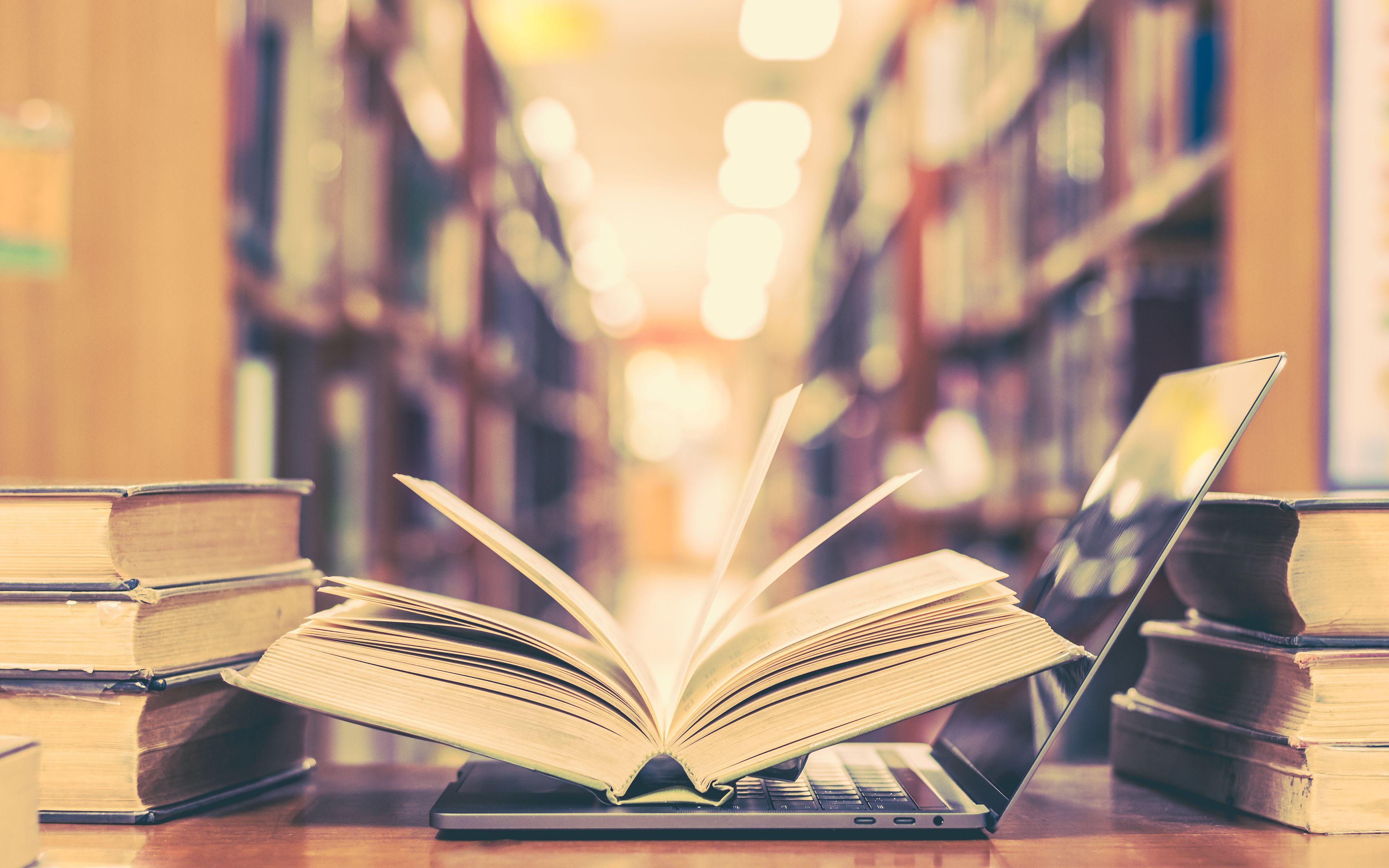 Between the shelves of a library are stacks of books and printed documents placed on top of a modern laptop.