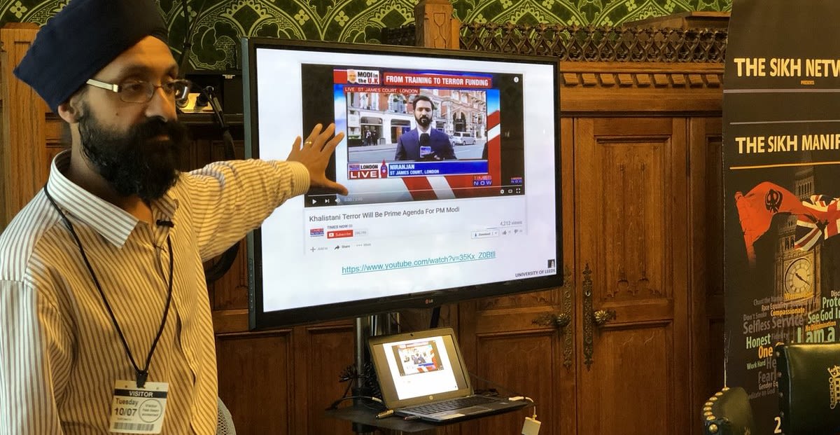 Dr Jas Singh pointing to a large tv or computer screen showing a news reporter