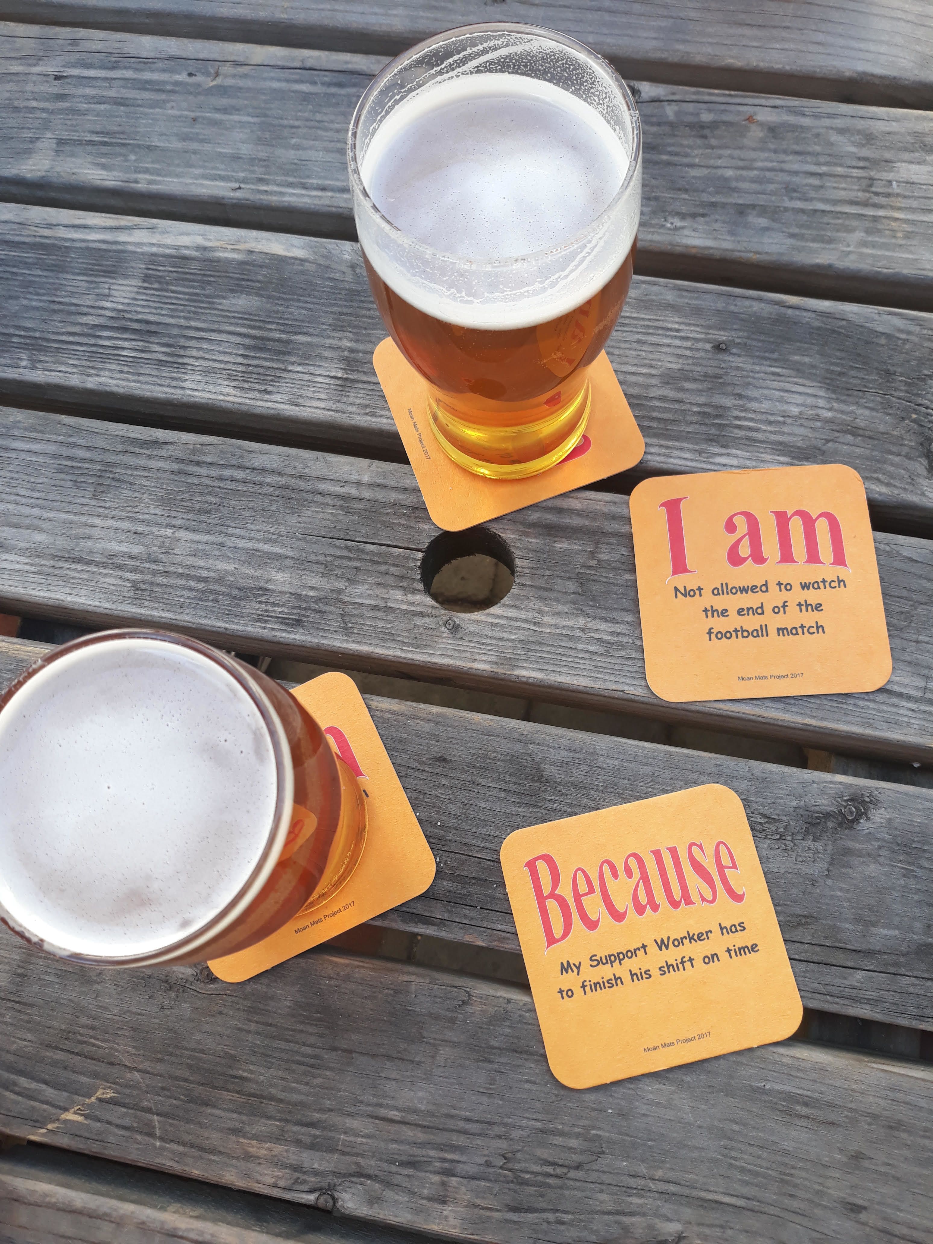 Pints of beer and Moan Mats by Stephen Harvey. The mats say: "I am not allowed the watch the end of the football match" on one side and on the reverse side, explains: "Because my support worker has to finish his shift on time."
